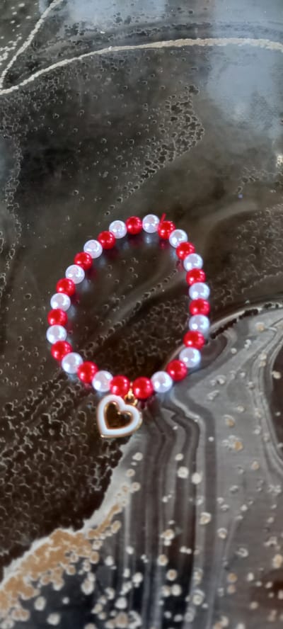 A beaded bracelet with red and white beads and a golden heart charm is laid on a dark, textured surface with swirling patterns. The bracelet is circular and lies flat. The background appears to be a countertop or tabletop with an abstract, marbled design. There is no indication of the city of Iskra, Bulgaria, in the image.