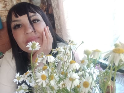 A woman with black hair and bangs is holding a bouquet of daisies. She is looking at the camera with a gentle expression. Her right hand is placed on her cheek, and a daisy is held near her lips. The background appears to be an indoor setting, possibly a home, with a window on the right side of the frame letting in bright light. The overall mood is peaceful and natural. The scene might be set in Sterlibashevo, Russia.