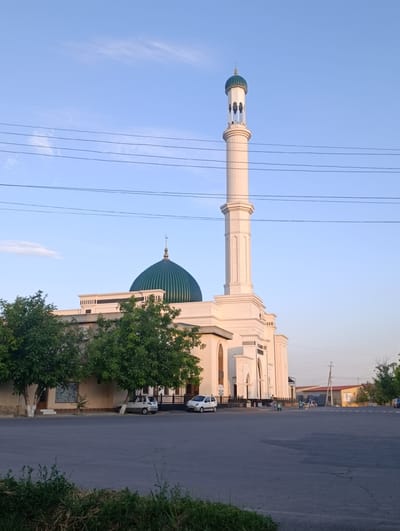 The image captures the exterior of a mosque situated in the city of Kibray, Uzbekistan. The mosque features a prominent white minaret topped with a green dome, and a large green dome on the main building. Several trees with lush green foliage are positioned in front of the mosque. A paved road runs in front of the building, with two white cars parked on the side. In the background, there are other buildings and utility poles, suggesting a semi-urban environment. The sky is clear blue with a few wispy clouds, and power lines are visible crisscrossing the upper portion of the image. The overall scene depicts a serene daytime view of a religious landmark within its local context.