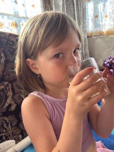 A young girl is shown from a close-up perspective, with the camera angled sideways. She is drinking from a clear glass with a yellow liquid inside, holding it with her right hand. She is wearing a pinkish-purple ribbed top. A small, purple sparkly object, possibly a toy or decoration, is visible near her face. Her hair is light brown and appears to be long, falling around her face. She has green eyes and is wearing a small red earring in her left ear. The background includes what appears to be a window with light blue curtains and a patterned brown blanket or rug. The lighting suggests it could be daytime, with sunlight illuminating the scene. The image is taken in Markidonovka, Russia.
