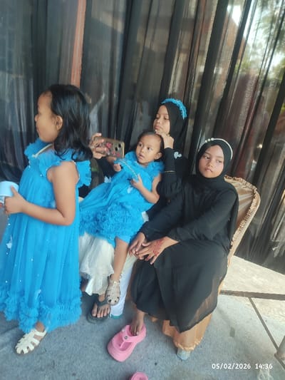 Four girls are posing together. Three of them wear matching blue dresses, and one of them uses a phone to take a photo of the youngest. One girl wears a black abaya with a white pearl headband, and another wears a matching black abaya with a blue headband. One girl wears white sandals, while the other wears pink flip-flops.