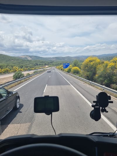 The image depicts a first-person perspective from the interior of a vehicle, looking forward through a windshield onto a multi-lane highway. The lower portion of the frame shows the vehicle's dark dashboard and part of a steering wheel. A black navigation device is mounted on the dashboard to the left of center, connected by a black cable. A black suction cup mount is visible on the dashboard to the right.
The highway features multiple lanes with white road markings; a solid white line defines the shoulder, and dashed white lines separate traffic lanes. A black SUV, possibly a Range Rover, is partially visible on the left, adjacent to the viewer's vehicle. Several other vehicles are discernible further down the highway in the same direction of travel, and on an elevated, parallel road to the left.
The surrounding environment consists of rolling hills extensively covered with green deciduous and coniferous trees, interspersed with numerous shrubs bearing vibrant yellow flowers. A blue highway sign, located on the right side of the road, displays the text "1000 m," an exit symbol, "N-232," and "Jérica (sur)." The sky is partly cloudy with significant white cloud cover and visible patches of blue sky. The scene is located on the N-232 road, approaching the southern exit for the city of Jérica in Spain.