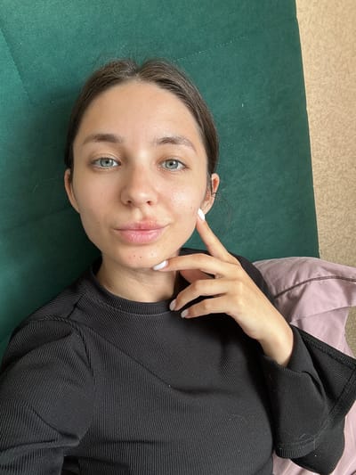 The media file is a selfie of a young woman lying on a dark green surface, possibly a couch or bed. She is looking directly at the camera with a neutral expression. Her head is tilted, and her right hand is raised, with her index finger touching her cheek near her eye. She is wearing a black, ribbed long-sleeved top. A pale pink pillow is visible behind her. The lighting suggests it is daytime, likely indoors. There are no visible text elements in the image. The setting appears to be a residential or casual environment.