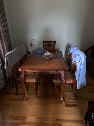 The image depicts a dining room setting in what appears to be a residential interior. The scene is captured from a slightly elevated perspective, with the camera oriented sideways. The primary objects in the frame are a polished wooden dining table and several chairs with upholstered seats and backs. One chair, positioned at the head of the table, has a light blue collared shirt draped over its back. On the table, a glass bowl filled with what looks like decorative items, possibly artificial fruit, is visible.

The room has wooden flooring with a distinct grain pattern. The walls are a neutral, light color. To the left of the table, a small, dark-framed mirror is mounted on the wall, and below it, a white electrical outlet with cables is visible. A light-colored towel or blanket is folded and placed on a darker surface, possibly a bench or the floor, in the lower left quadrant of the image. In the upper right corner, a portion of a dark brown blanket with an embroidered design is visible.

The lighting in the room appears to be artificial, casting shadows and highlighting the wood grain and the sheen of the table. There is no indication of the time of day or weather conditions. The scene suggests a quiet, indoor environment, possibly a home. No people are visible in the image. There is no visible text in the frame. The overall impression is one of domestic comfort and order, with the draped shirt being the only element suggesting recent human presence or preparation for an activity.