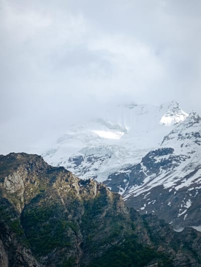 The image depicts a majestic mountain range, with a dramatic contrast between the rocky, verdant slopes in the foreground and the snow-capped peaks in the background. The sky above is overcast with soft, diffused light, contributing to a serene and slightly dramatic atmosphere. The lower portion of the mountains is covered in dark green vegetation, suggesting a temperate climate or a lower altitude. The upper reaches are dramatically covered in snow and ice, with visible glacial features and sharp, rocky outcrops peeking through. The lighting highlights the textures of the rock and the contours of the snow, creating a sense of depth and grandeur. The scene appears to be a natural landscape, devoid of any human presence or artificial structures. The composition, with the foreground mountains leading the eye towards the imposing snow-covered giants, emphasizes the scale and wildness of the environment. This vista is consistent with the dramatic mountain scenery found in regions like Gemur, India, known for its high-altitude terrain and glacial landscapes.