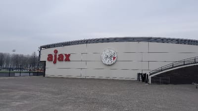 The image displays the exterior of a building associated with AFC Ajax, situated in Amsterdam-Duivendrecht, Netherlands. The structure, constructed with light grey horizontal wall panels, features large, red "ajax" lettering prominently on its left side. To the right of the lettering, a circular white emblem depicting the stylized head of the Greek hero Ajax with a red accent is affixed to the wall. A dark grey, curved external staircase with black handrails ascends along the right side of the building. Below the staircase, a gated, dark recessed entrance is visible. The foreground consists of a uniform grey paved surface. In the background to the left, bare deciduous trees, a tall light standard, and a section of a fenced sports field are observable. The sky is uniformly overcast. No individuals or active subjects are present in the scene.