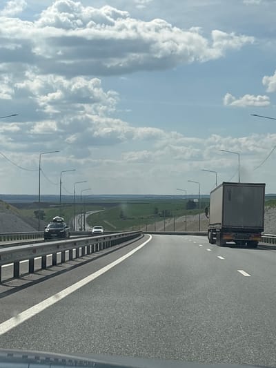 A gray truck travels on a highway under a cloudy sky. The truck is on the right side of the road, heading away from the viewer. To the left of the truck, there is a white car and a black SUV with a roof rack. The highway is flanked by a guardrail and a grassy embankment. In the distance, there are rolling green hills and a body of water. The scene appears to be in the city of Asanovo, Russia, though this is not explicitly visible in the image.
