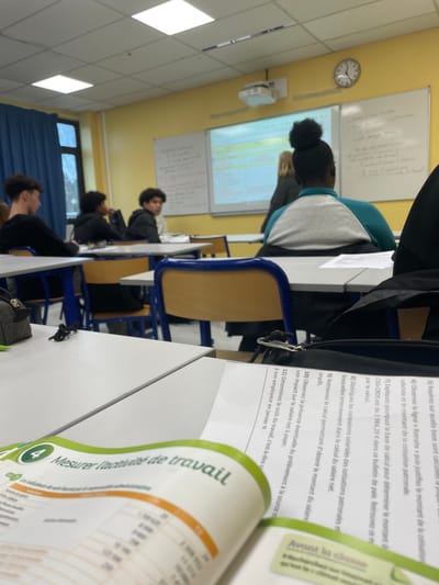 This image depicts the interior of a classroom within a secondary educational institution in Hérouville-Saint-Clair, France. The perspective is from a student's desk in the foreground, looking towards the front of the room and diagonally across other seated students.
In the immediate foreground, an open textbook rests on a white desk. Its visible text, primarily in French, includes the chapter title "Mesurer l'activité de travail" (Measure the activity of work) and questions such as "Déterminez le coût du travail" (Determine the cost of labor). A black zippered bag or pencil case is partially visible on the right side of the foreground desk.
Multiple students are seated at individual white desks with blue and light brown chairs. In the mid-ground, a student with dark, curly hair styled in a bun is seated, facing forward, wearing a grey and teal colored sweatshirt. To the left and further back, a male student with dark hair, dressed in a dark long-sleeved shirt, is seen in profile. Adjacent to him, another male student with dark hair and skin faces the front. Behind these individuals, a male student with curly dark hair, wearing a dark jacket, is seated and appears to be looking towards the camera's position.
The classroom features yellow-painted walls and a ceiling with rectangular recessed light fixtures. Two whiteboards are mounted on the front wall. A white multimedia projector is suspended from the ceiling, projecting illuminated content onto the left whiteboard. A circular analog clock, displaying approximately 10:45, is mounted on the yellow wall above the right whiteboard. A large window with blue curtains is visible on the left side of the room, showing an indistinct outdoor view.