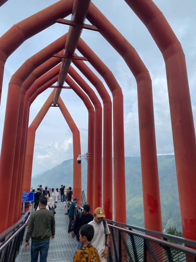 The image depicts the Skywalk Turpuk Malau in Turpuk Malau, Indonesia. A group of people are walking on a metal walkway lined with bright orange, arched tubular structures. The walkway has a railing on the sides. In the background, lush green mountains and valleys are visible under a cloudy sky. There is a sign on the left side of the walkway. A security camera is mounted on one of the orange structures. The scene appears to be a tourist attraction, with visitors enjoying the panoramic views.