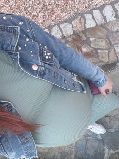 The image, taken from a high angle, shows a person standing on a stone surface. The person is wearing a light green, ribbed dress and a blue denim jacket adorned with pearls. They are holding a small, purple wallet in their left hand. A white sneaker is visible near the right edge of the frame. Reddish-brown hair is visible near the bottom right corner. The background consists of a light-colored stone surface. The location and specifics related to the city of Brusen, Bulgaria, are not discernible from the image.