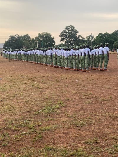 The image displays approximately 100 individuals, presumed to be young adults, standing in multiple structured rows on a reddish-brown earthen field with sparse green vegetation. Each person wears a uniform consisting of a white short-sleeved shirt, light green/khaki trousers, a matching cap, and light brown/tan boots. The shirts are marked with "NYSC" on the back. The individuals are in a formal formation, facing towards the left of the frame. In the background, a line of dense green trees separates the field from distant structures and a visible green and white flag. The sky is overcast. This scene is located in Kubi A, Nigeria.