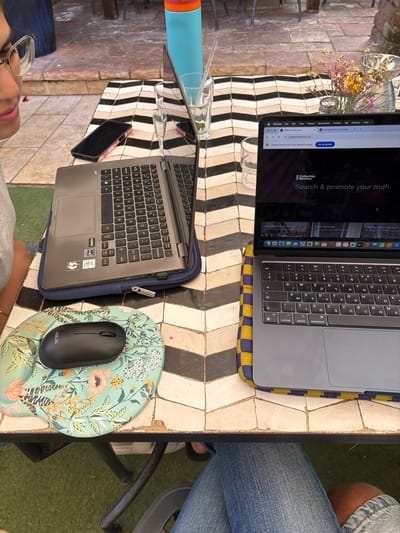 A table with a black and white checkered tile pattern is positioned outside. Two laptops are open and positioned on the table. One laptop is a black and silver Acer and the other laptop is a silver and black Apple laptop.  A blue and floral patterned mouse pad sits on the table to the left of the Acer laptop.  A black computer mouse sits on top of the mouse pad.  There is a blue and floral patterned smartphone on the table to the left of the Acer laptop.  A person's hand holding a blue water bottle sits outside of the frame.  A pair of legs, one clothed in blue jeans and the other in khaki pants, sit at the table.  The table is in front of a brick wall and a green grassy area.  The location of the table is at the coordinates 32.173, 34.872 in the city of Ra'anana, Israel.