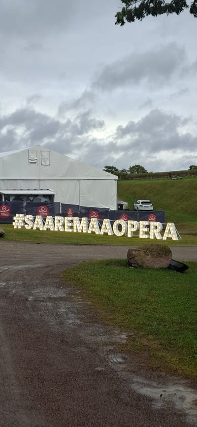 This image shows an outdoor event setup in Kuressaare, Estonia. The main subjects are a large white tent, a prominent lighted sign spelling "#SAAREMAAOPERA", and a natural landscape featuring a grassy hill and a dirt path. The sign, made of illuminated individual letters, stands vertically and is a focal point. Behind the sign, a large, moss-covered rock rests on the grass.
The scene appears to be during the daytime, under a cloudy, overcast sky, suggesting potentially damp weather, as evidenced by the wet appearance of the dirt path. A white SUV is parked in the background near the tent. The tent itself is large and white, with some visible windows or openings and signage on its side. The overall atmosphere is functional and preparatory for an event, with no people visibly present in this particular frame.
The text clearly visible is "#SAAREMAAOPERA" on the illuminated sign. The presence of the tent and the dedicated signage suggests an outdoor performance or festival, likely related to opera, taking place in Saaremaa, Estonia. The natural setting of the grassy hill and the rock contribute to the outdoor ambiance.