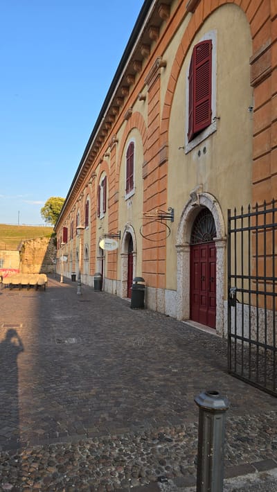 The image captures a street scene in Peschiera del Garda, Italy, with a focus on the facade of a long, historic building. The building is painted a pale yellow with prominent orange-red brick arches framing windows and doorways. Red shutters are visible on some of the windows. The street is paved with cobblestones, and a dark metal gate is positioned on the right side of the frame. Several dark trash receptacles are placed along the building's exterior. In the background, a grassy embankment with a tree can be seen, suggesting the building is situated near a park or fortified area. The lighting indicates it is either early morning or late afternoon, with long shadows cast across the cobblestones, including the shadow of a person holding a phone. Chairs are stacked in the distance on the left side of the image. A sign for "Rotary Villafranca" is attached to the building's facade.