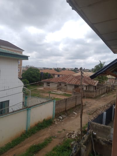 The image is an outdoor, eye-level shot looking across a neighborhood with a cloudy sky. On the left, a large, modern building with white tiled walls and green trim is visible. In the center and right of the image are several smaller, older buildings with corrugated metal roofs, surrounded by a mix of dirt paths and patches of grass. A tall, thin wooden pole with electrical wires is a prominent feature in the foreground. Barbed wire fences line some of the property boundaries. In the background, more buildings with red-tiled roofs and palm trees can be seen, suggesting a semi-urban or residential area. The overall impression is of a typical street scene in a warm climate.