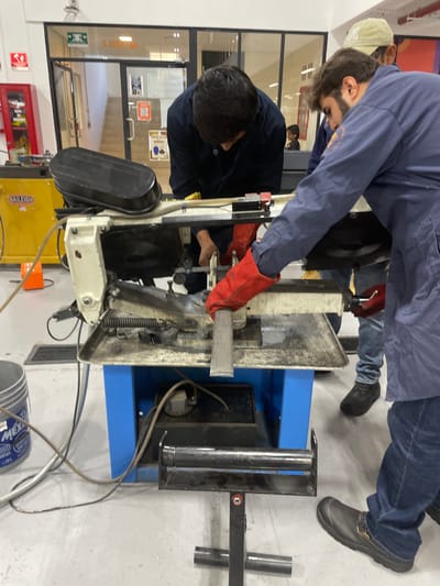 The scene depicts three individuals engaged in metalworking within a workshop environment located in Fraccionamiento Los Heroes Queretaro, Mexico. The primary focus is a beige and black horizontal bandsaw machine, mounted on a blue stand, actively cutting a rectangular metallic bar. A male individual, positioned on the right, wears a blue long-sleeved work coat and red protective gloves, with his right hand extended towards the cutting area of the machine. Another male, positioned to the left of the machine, wears a dark blue long-sleeved shirt and appears to be observing or assisting, with his hands near the machine's controls. A third male, partially visible behind the first, wears a light-colored baseball cap and blue jeans. The bandsaw's cutting area shows evidence of lubricant or coolant, with residue on the machine's tray.
In the background, a set of glass double doors with "ACCESO S 02" (likely mirrored) and an emergency exit sign are visible, revealing a light-colored staircase beyond. To the far left, a red cabinet labeled "CASO DE INCENDIO" (Fire Case) and yellow industrial machinery, partially labeled "BAILEIGH," are present. An orange safety cone is on the floor near the yellow machinery. A silver bucket with "MEXICO" and "DIESEL" labeling is on the floor to the immediate left of the bandsaw. Various electrical cords and hoses connect to the machinery. The floor is light gray concrete. A black metal roller stand is situated on the floor in front of the bandsaw. A reflection of a fourth individual, possibly female, is faintly visible in the glass door.