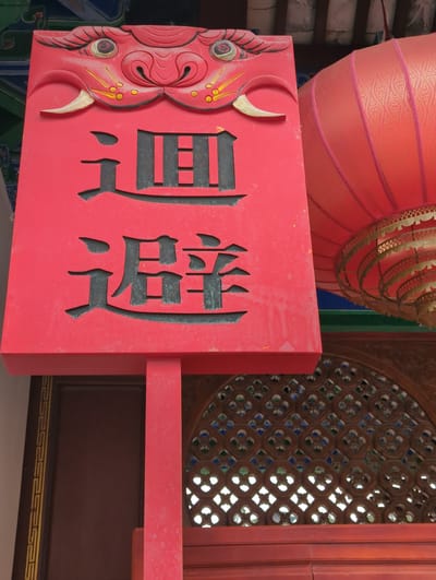 This is a photograph of a red wooden sign with black Chinese characters carved into it. The sign is adorned with a red, dragon-like motif on the left side, featuring carved eyes and a horn. The Chinese characters appear to read "景觀" (jǐngguān), meaning "scenery" or "view."
Behind the sign, a large, ornate red lantern with gold embellishments hangs from above. To the right of the sign, a wooden lattice window with intricate geometric patterns and colored glass inserts is visible. The overall setting suggests a traditional Chinese architectural environment, possibly a temple, garden, or a historic building. The red color of the sign and lantern, combined with the gold and intricate designs, evokes a sense of cultural richness and celebration. The lighting suggests it is daytime, with natural light illuminating the scene. The photograph is taken at a close-up angle, focusing on the sign and its immediate surroundings. There is no visible indication of people or a specific event taking place, but the elements present suggest a place of cultural significance or historical interest in Gucheng District, China.