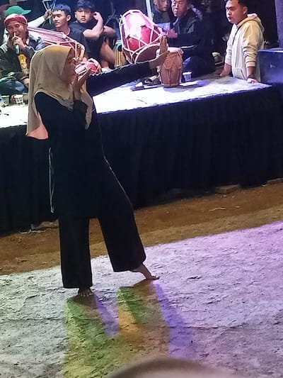 The image captures a traditional cultural performance, likely in the evening or at night, in Pangalengan, Indonesia. The central subject is a woman, possibly a dancer, wearing a light-colored hijab, a dark long-sleeved tunic, and dark wide-leg pants. She is barefoot, actively performing a dance move, balancing on one foot with the other's toes touching the ground behind her. Her left arm is extended forward, holding a traditional red and white drum (kendang) upright. Her right hand is positioned near her mouth.
In the background, several men are present around a long table draped with dark fabric. One man on the far left, wearing a green cap, holds a microphone, suggesting he is singing or speaking. Other male figures, including one in a light-colored hoodie on the right, are observing or participating in the musical ensemble, with another smaller drum visible on the table. The ground where the woman dances appears to be packed dirt or concrete, illuminated by artificial colored lights that cast vibrant purple, green, and yellow reflections. The background beyond the performers is dark, highlighting the stage area. The scene depicts a focused cultural presentation involving music and dance. No visible text is present.