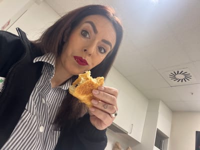 A woman is holding a partially eaten pastry in her hand, with a ring on her finger that features a small heart tattoo. She is wearing a striped shirt under a dark jacket. The location appears to be indoors with a white, tiled ceiling featuring a circular vent. White cabinets are visible in the background. There is no indication of the city, Mensdorf, Luxembourg in the image.