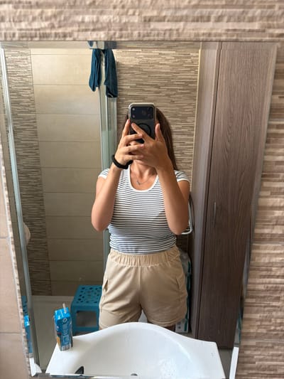 A young woman stands in a bathroom, taking a mirror selfie with her phone. She is wearing a striped t-shirt and beige shorts. The bathroom features tiled walls, a wooden cabinet, a sink, and a shower. A blue stool and a juice box are visible on the counter. The scene appears to be in a private residence in Dromolaxia - Meneou Municipality, Cyprus.