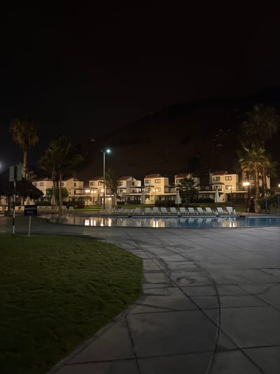 The image depicts a nighttime scene at a resort or hotel complex, likely in San Antonio, Peru, given the context. A swimming pool with illuminated reflections dominates the foreground. Beyond the pool, a row of white buildings with red-tiled roofs are visible, featuring balconies and illuminated windows. Palm trees are scattered throughout the property, and lounge chairs are arranged around the pool area. A grassy area is to the left of the pool, and a sign is visible near the left edge of the frame. The background features a dark, hilly landscape. The overall impression is one of a tranquil, illuminated resort at night.