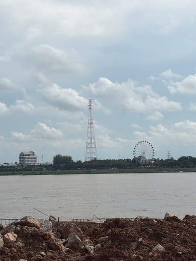 The image captures a wide-angle view of the Mekong River in Thakhek, Laos, on a cloudy day. In the foreground, a pile of brown soil and rocks is visible, suggesting ongoing construction or landscaping. The river itself occupies a significant portion of the lower half of the frame, its surface appearing murky and rippled.
Across the river, the opposite bank is lined with lush green trees. Dominating the middle ground of the landscape are several man-made structures. To the left, a large, modern white building with multiple floors stands out. Further to the right, a tall, red and white lattice tower, likely a transmission tower, is prominently featured. In the distance, a large Ferris wheel, a common feature of amusement parks, is visible, adding a touch of entertainment to the scene. The sky above is filled with a mix of white and grey clouds, indicating overcast weather. The overall impression is one of a developing urban area situated along a major river.