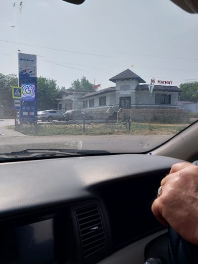 A view from inside a car shows a building with a sign that reads "МАГНИТ" in Russian, indicating a Magnit store. To the left of the Magnit store, a tall sign displays "МЕГАПОЛИТ" in Russian, suggesting a connection to a gas station or service center. Below the "МЕГАПОЛИТ" sign, a smaller blue sign shows a green "G" logo with the words "КАЧЕСТВО ПРОВЕРЕНО" and "ГАЗПРОМНЕФТЬ" which translates to "QUALITY CHECKED" and "GAZPROMSERVICE", further confirming a gas station. There are also pedestrian crossing and bus stop signs visible. The scene appears to be on a street in Belokurikha, Russia, with trees and a cloudy sky in the background. The interior of the car is visible, including the dashboard and a person's hand on the steering wheel.