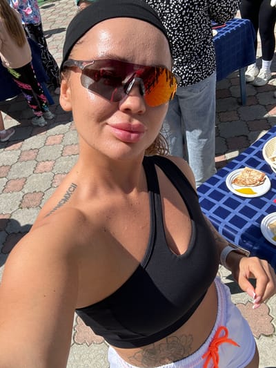 A woman with a bandana and sunglasses takes a selfie outdoors. She is wearing a black sports bra and white shorts with an orange drawstring. Her left shoulder has a tattoo of Roman numerals. In the background, people are standing near tables with food, including pancakes and honey. The ground is paved with patterned tiles. The scene appears to be in Duvankul', Russia.