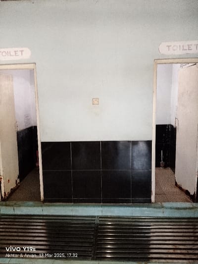 The image shows a view of two toilet stalls. The stalls are located in a building, possibly a public restroom or a public facility. The stall on the left has a sign above it that reads "TOILET," while the stall on the right also has a sign that reads "TOILET." Both stall doors are open, and there is a small, square electrical outlet visible on the wall. The stalls are lined with black tiles, and a light blue tile trim separates the black tiles from the pale blue wall. There is a stainless-steel drainage grate in the floor in the middle of the image. The image is taken from a slightly elevated perspective. The location of the image is in Kecamatan Gabuswetan, Indonesia, at latitude -6.44 and longitude 108.027. There are no people visible in the image.