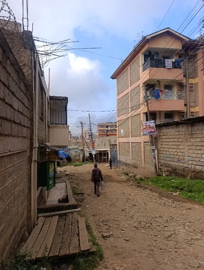 An unpaved, rocky street scene depicts a low-income neighborhood. Buildings made of brick and concrete flank the street, some topped with barbed wire. Overhead, power lines crisscross the sky. A person walks away from the viewer, while another figure stands farther down the road. Wooden planks sit beside a wall on the left, and patches of grass dot the edges of the street. Laundry hangs on a balcony of one of the buildings.