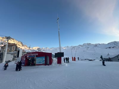 This image depicts a snow-covered ski slope at the Val Thorens ski resort in Saint-Martin-de-Belleville, France, under a clear blue sky with distant snow-capped mountains. In the mid-ground, a prominent red building marked with "Val Thorens" signage and a large ski map display appears to be a ski lift entrance or information point. To the left of this building, a large grey ski lift pylon supports cables with several chairs ascending and descending. Further left, a multi-story building complex is partially visible. A tall, slender grey pole with attached sensors or lights stands to the right of the red building. Approximately ten to fifteen individuals are present on the snow. Several people in winter attire are gathered near the red building, while others are seen moving across the slope. One person on the far right is actively snowboarding, facing left. The snow-covered ground shows minimal tracks.