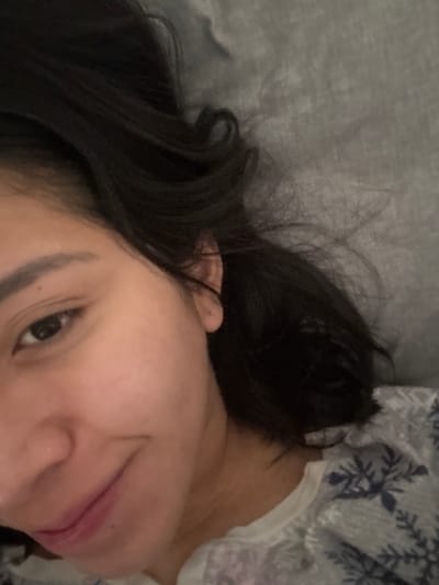 The image displays a close-up view of an individual, appearing female, in a reclined position. The subject's face is partially visible, featuring the left eye directly engaging the camera and a subtle smile on the visible portion of the lips. The skin tone is light to medium. Dark, medium-length hair is spread across a plain, uniform gray background, consistent with a pillow or bed linen. The individual is wearing a white garment patterned with blue or gray snowflake-like designs on the visible upper torso. The lighting is soft and even, indicating an indoor environment. No specific visual elements within the image serve to confirm the stated location of Atizapán de Zaragoza, Mexico.