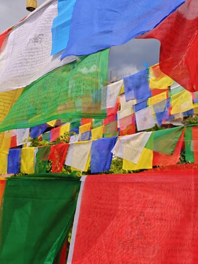 The image displays numerous prayer flags in various colors, predominantly red, blue, green, and white, arranged in multiple layers. Some flags bear inscriptions and imagery. The flags are suspended outdoors, with a cloudy sky visible in the background. Green foliage is also present, partially obscured by the flags. The scene appears to be located in Gokarneshwar, Nepal, a site often adorned with such prayer flags, typically near religious or spiritual locations. There are no people or animals visible in the image.