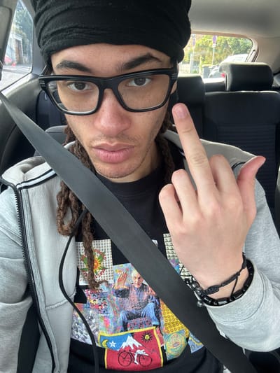A man is seated in what appears to be a car, wearing a seatbelt. He is wearing a black beanie, glasses, and a grey zippered hoodie over a black t-shirt with a colorful graphic print. His hair appears to be styled in dreadlocks or braids. He's holding his hand up, showing his index finger. The car's interior and a glimpse of the outside scenery through the window are visible. The image was taken in the city of Orsay, France.