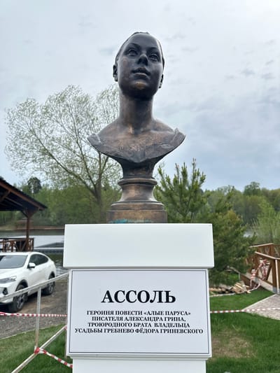 The image shows a bronze bust of Assol, the main character in Alexander Grin's novel "Scarlet Sails," located in Grebnevo, Russia. The bust is mounted on a pillar, next to a sign that identifies Assol as the heroine of the novel and mentions the connection between the author and the owner of the Grebnevo estate. The surroundings include trees, grass, a wooden structure, a car, and a red-and-white striped barrier tape. The sky is overcast.