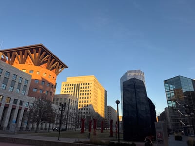 An urban landscape under a clear sky showcases a variety of architectural styles. A reddish-brown building with a distinctive overhanging roof structure stands prominently beside a gray building with vertical columns. In the background, a light-colored, block-shaped building basks in sunlight. A dark, abstract sculpture is juxtaposed with a modern glass building. Bare trees line a grassy area, accented by small, red vertical structures. The scene is bathed in the warm, early light of the day.