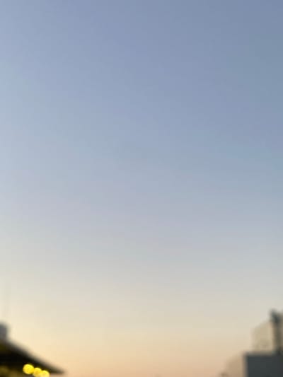 This is a medium shot, taken outdoors, capturing a hazy sky during what appears to be either sunrise or sunset. The vast majority of the frame is filled with a gradient of pale blue transitioning to a soft, warm yellow on the right side, suggesting the presence of the sun just below the horizon or its lingering glow.

In the upper right corner, partially obscured by the blur and haze, are faint outlines of buildings. One structure has a darker, possibly grey, section, and what looks like a window or opening. Lower down on the right, a cluster of out-of-focus yellow lights, resembling fairy lights or small lanterns, are visible. Their bokeh effect further emphasizes the soft focus of the scene.

The overall impression is one of a calm and serene moment, possibly on a slightly overcast day given the diffused light. The lack of sharp details and the gentle color palette contribute to a peaceful ambiance. There is no visible text in the image. The location context provided is Bolesławiec, Poland.