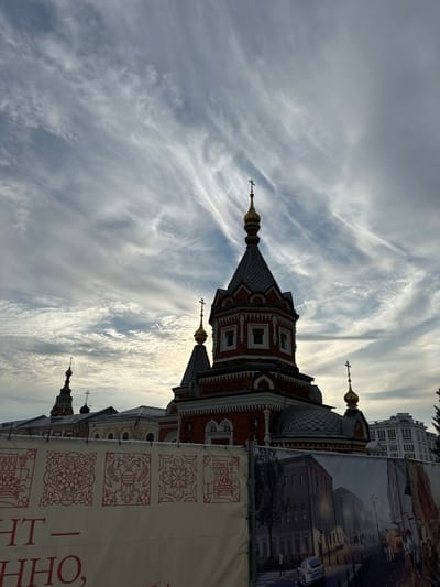 The image shows an Orthodox church with multiple domes, silhouetted against a cloudy sky. A banner with decorative patterns and text in Cyrillic characters is in the foreground, obscuring the lower portion of the church and surrounding buildings. The banner also features a printed image of a street scene with buildings and people. The overall impression is that of an urban environment, possibly undergoing some form of construction or renovation given the presence of the banner. The location is identified as Putyatino (Semivragovskaya S/A), Russia.