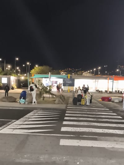 The image depicts an outdoor scene at night, likely an airport or transportation hub, in Sainte-Marie, France. A group of people are visible, some walking with luggage across a pedestrian crossing. The environment includes a building with illuminated windows, streetlights, trees, and large rocks. The lighting suggests it is dark, with artificial lights illuminating the area. Several individuals are pulling or pushing luggage carts and suitcases. The pedestrian crossing is marked with white stripes on the asphalt. In the background, a cityscape with lights is visible on a hill.