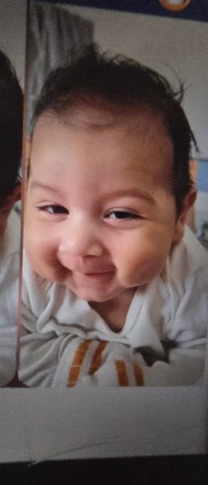 The image depicts a close-up portrait of an infant, situated in a presumed indoor location within Giengen an der Brenz, Germany. The infant, characterized by dark hair and light brown skin, is positioned on its stomach, with its head lifted and face directed towards the viewer. It is wearing a light-colored, long-sleeved garment. Beneath the infant's chin, a fabric with distinct vertical orange or yellow stripes is visible. The infant's facial expression is a wide, open-mouthed smile, with eyes partially closed due to the smiling action and raised cheeks. The background elements are out of focus, revealing indistinct patterns and colors consistent with an indoor environment. A partial view of another individual's head or dark hair is visible on the far left edge of the frame.