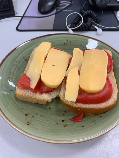 The image displays two open-faced sandwiches on a speckled green plate, viewed from directly above. Each sandwich consists of a slice of bread topped with slices of red tomato and slices of yellow cheese. One sandwich has two slices of cheese, one larger and more curved, and a smaller, thinner slice underneath. The other sandwich also has two slices of cheese layered on top of the tomato. The bread appears to be a light-colored, possibly white or sourdough bread. The tomatoes are bright red and appear to be sliced fairly thickly. The cheese is a solid yellow and has a smooth texture, with one slice showing a small, circular hole. A small piece of red tomato is visible on the plate to the right of the sandwiches.
In the background, to the left of the plate, is a dark surface with a mouse and a keyboard visible. A coiled black cord and a white earbud wire are also present on this surface, along with white earbuds. The overall setting suggests an indoor environment, possibly a desk or office space, given the presence of the computer peripherals. The lighting is bright, indicating it is daytime. There is no visible text in the image. The activity taking place is the preparation or consumption of food.