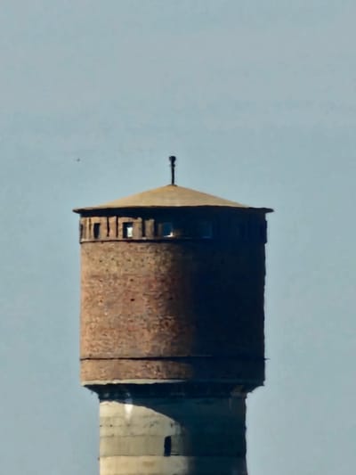 A tall, brick water tower stands against a pale blue sky. The tower has a circular base and a cylindrical upper section constructed of weathered, reddish-brown bricks. The top of the tower is capped with a conical roof, possibly made of metal, and a small, thin antenna or spire protrudes from the apex. A band of small, rectangular windows encircles the upper part of the brick section, just below the roofline. The lower portion of the tower appears to be made of concrete, with visible structural elements and a darker, arched opening. The overall impression is of an old, functional industrial structure. The image is a close-up view, framing the upper and middle sections of the tower against the expansive sky.