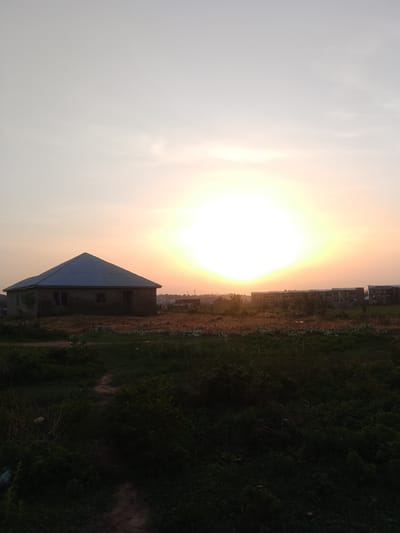 The image shows a rural scene at dusk or dawn in Ibo, Nigeria. A small, unpainted, single-story house with a corrugated metal roof stands in a field of low-lying vegetation. The sun is positioned low in the sky, creating a bright, hazy glow that obscures details in the middle distance. In the background, there are unfinished or partially constructed buildings. The foreground is dominated by dark green vegetation and a dirt path.