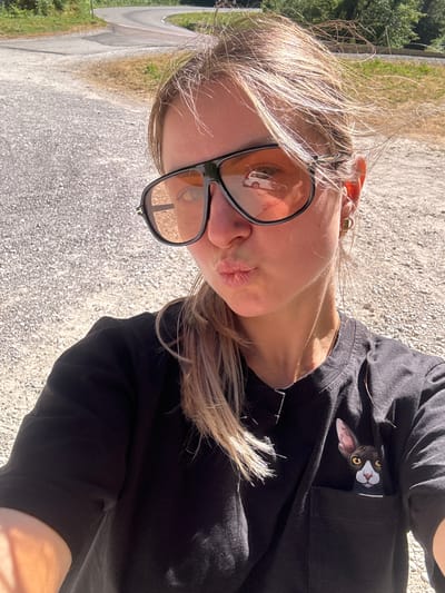 This is a selfie of a woman wearing sunglasses and a black t-shirt with a cat's face printed on the pocket. The woman is outdoors with a winding road and trees in the background, possibly in L'Essard, France. The sun is shining, creating shadows and highlighting the woman's hair and facial features.