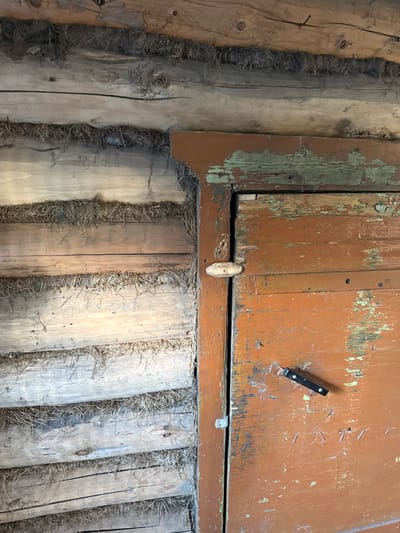 An exterior close-up view shows the corner of a rustic log cabin construction, with horizontally stacked logs chinked with dried grass. The right side of the frame features a weathered, orange-painted wooden door, with visible peeling paint revealing green undertones. A simple wooden latch secures the door from the outside, and a small black object, possibly a keychain or a tool, is attached to the door with two small screws. The overall scene suggests an aged and utilitarian structure, typical of rural or historical dwellings. The location of Чуй-Атасево, Russia, is associated with this structure.