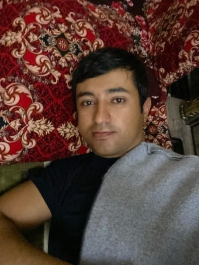 A man lies on a patterned red and beige textile. He has short, dark hair and brown eyes. He is partially covered by a gray garment. The man wears a dark blue or black shirt. The image location is Galaosiyo, Uzbekistan.