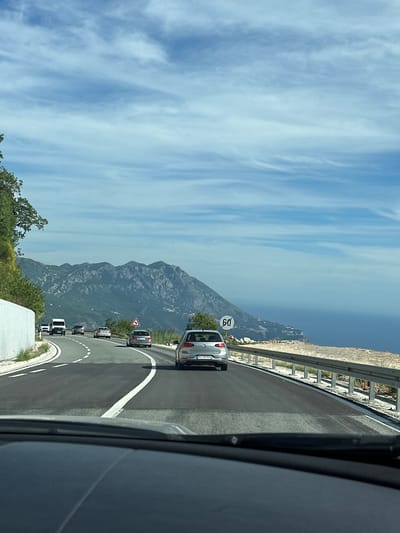 The image shows a view from inside a car driving on a winding road. The road curves to the right, with a white concrete wall on the left and a metal guardrail on the right. Beyond the guardrail, there is a steep drop to the sea, with a coastal town visible in the distance. Mountains rise behind the town. Several cars are on the road, and a speed limit sign indicating "60" is visible on the right. The sky is blue with scattered white clouds. The scene appears to be in a mountainous coastal region, likely Vrela, Montenegro, given the presence of a winding road, the sea, and dramatic mountain topography.