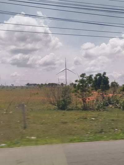 The image depicts a landscape with wind turbines in the distance, under a cloudy sky. The foreground shows a grassy area with a dirt path and some trees. Power lines are visible across the top of the image. The scene appears to be a rural or semi-rural area. While the prompt mentions the city of Hòa Thắng, Vietnam, the image itself does not contain any specific visual cues that would allow for precise location identification within that city. The overall impression is one of an open, windy landscape, likely a wind farm.