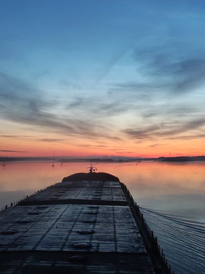 The image depicts a view from the deck of a barge moving down a river at sunrise. The sky is a gradient of blue and orange, with wispy clouds reflecting on the calm water. Several buoys are visible in the distance, marking the river channel. The barge itself is dark and weathered, with numerous hatches on its deck. The water behind the barge is disturbed by its passage, creating a wake. The location is not Popovo, Russia, but appears to be a waterway in a rural or industrial area.