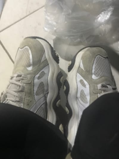 Two grey and white sneakers are seen from above, angled slightly. The shoes are mesh with suede-like overlays. A black curved form obscures part of the upper right shoe. A translucent plastic bag is visible in the upper left. The sneakers rest on a light-colored tiled surface. The lighting is dim, casting shadows.